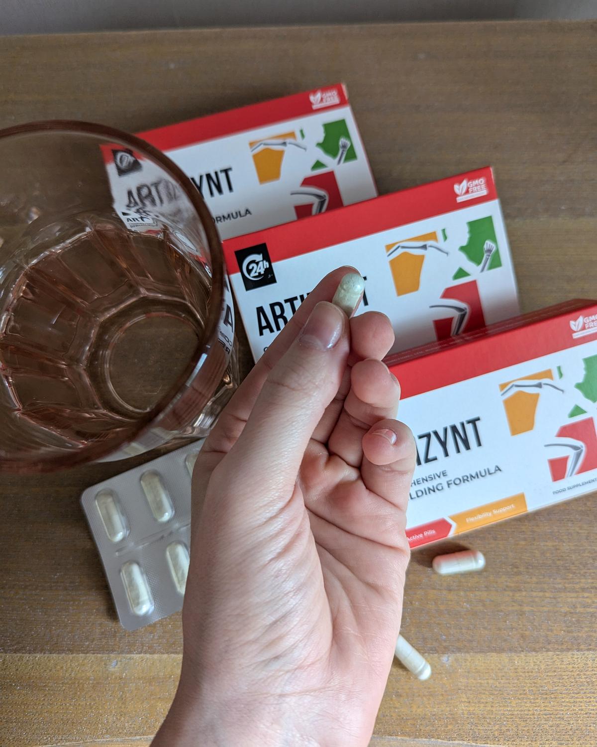 Hand hält eine weiße ArtiZynt-Kapsel über den Packungen, daneben ein Glas Wasser und ein Blister